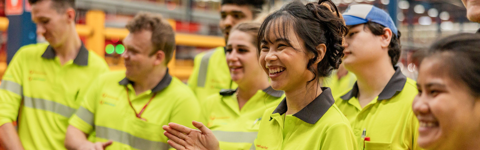 Retail team member in uniform smiles to camera; Warehouse team member in uniform smiles to camera, Office team member in a colourful dress smiles with coffee and notebook, Truck Driver in uniform talks to team member.