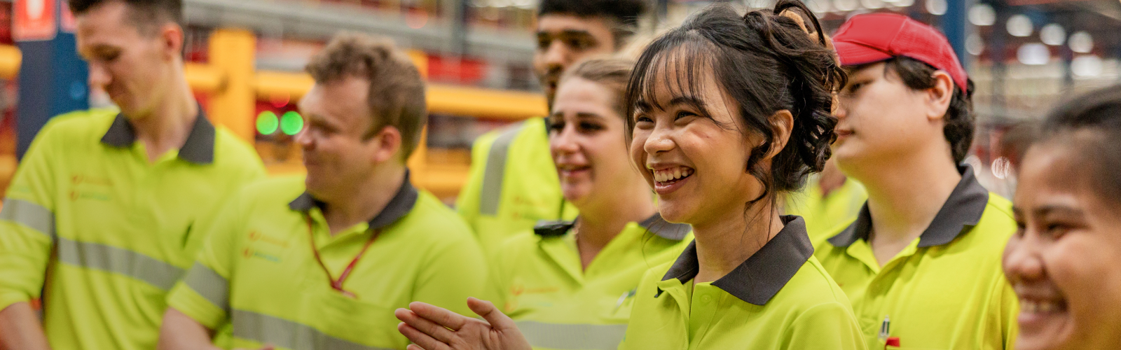 Warehouse team member in hi-vis uniform smiles and claps at person speaking off camera. Other coworkers in background are watching and listening to the person speak off camera.