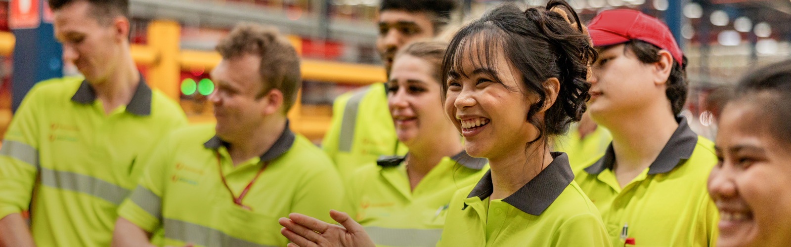 Retail team member in uniform smiles to camera; Warehouse team member in uniform smiles to camera, Office team member in a colourful dress smiles with coffee and notebook, Truck Driver in uniform talks to team member.