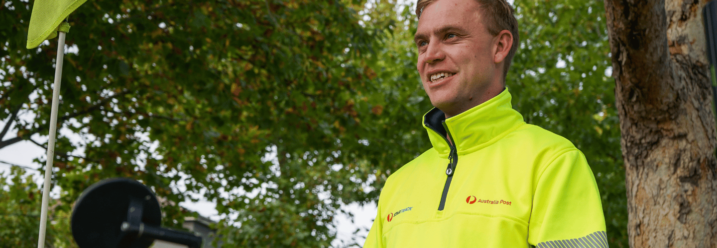 Postie in hi-vis jacket standing outside with trees behind him. 
