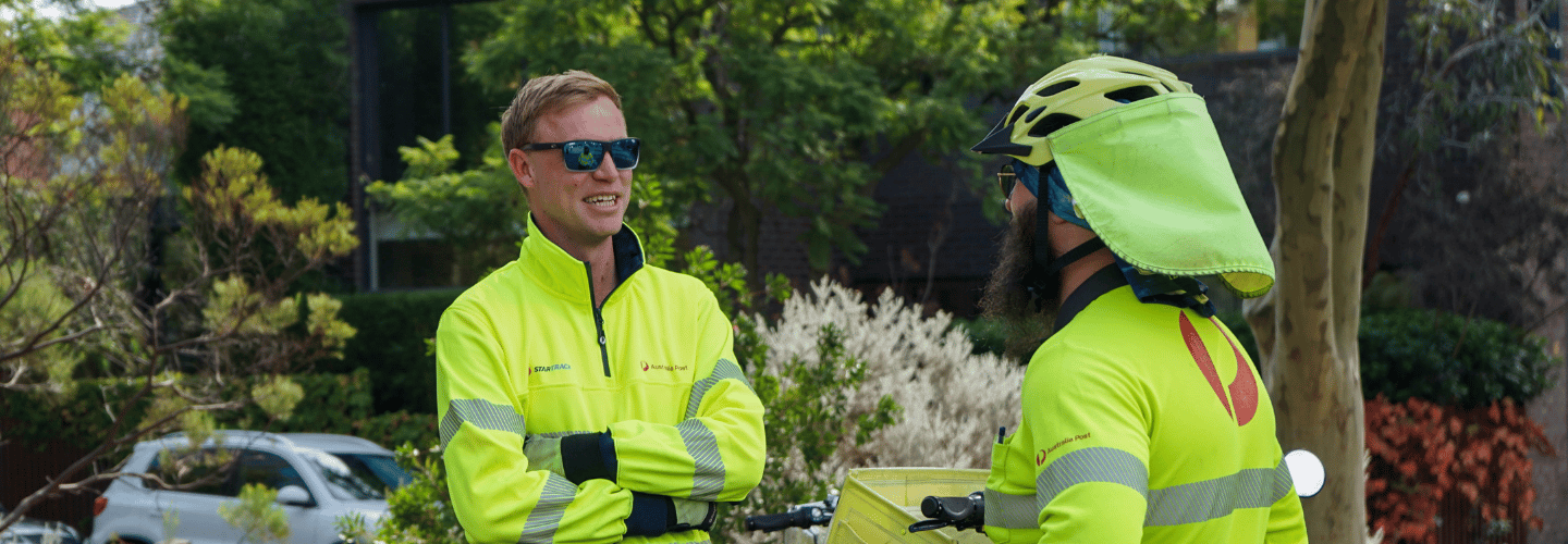Two Posties stand outside chatting and smiling while on a break in a park
