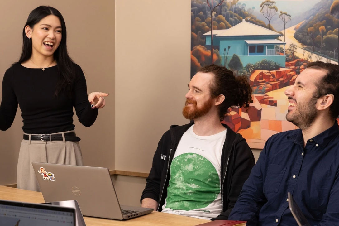 Two office team members stand chatting and laughing by their desk.