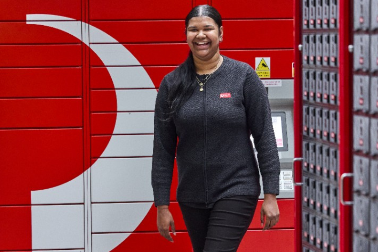 A retail team member smiles, walking past Postal Lockers and Post Boxes.