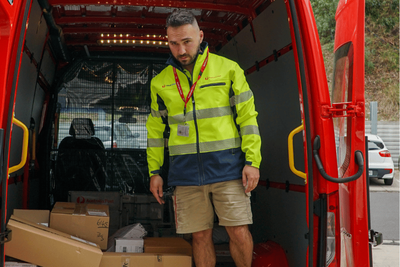 Van driver stands inside back of van filled with parcels and looks into camera 