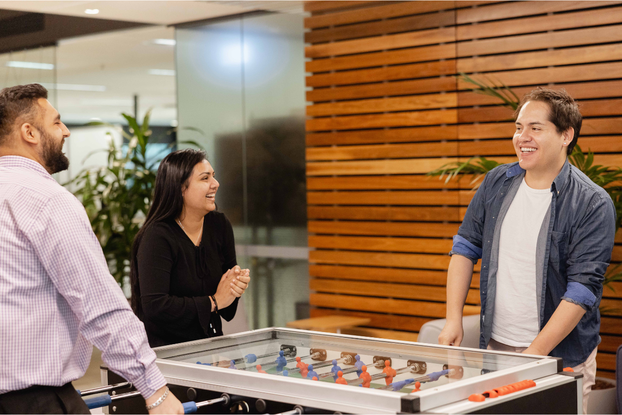 Three team members play at a foosball table together laughing.