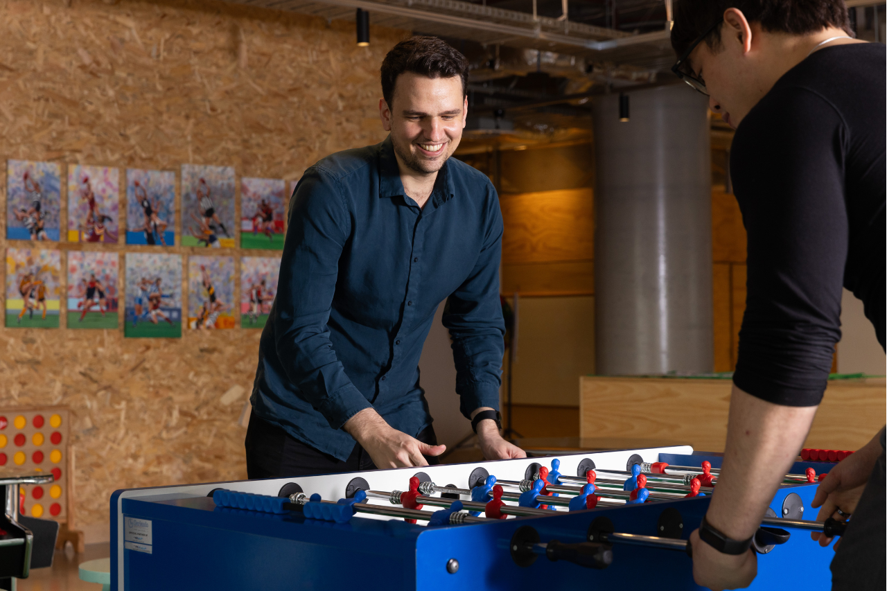 Three team members play at a foosball table together laughing.