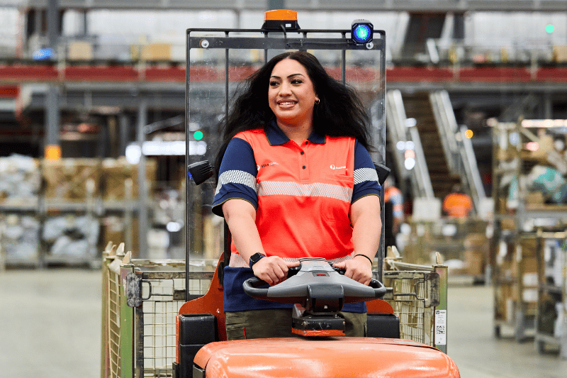 Warehouse team member driving a tugger across facility floor with facility in background. 